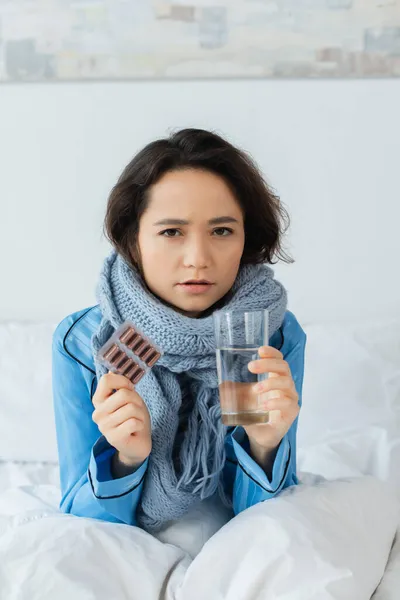 Sick woman in knitted scarf holding glass of water and blister pack with capsules — Stock Photo