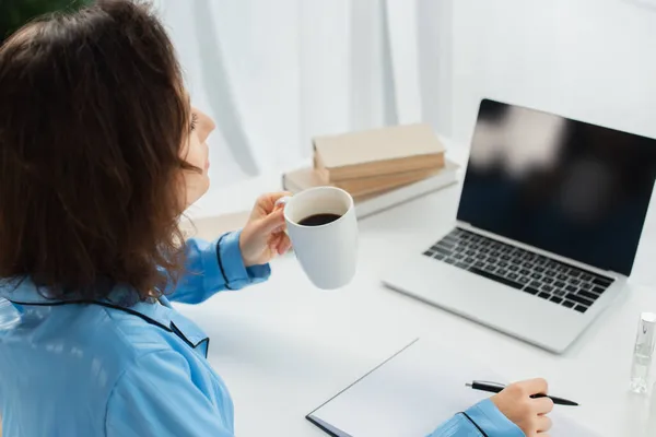 Mulher morena segurando xícara de café perto de laptop com tela em branco na mesa — Fotografia de Stock