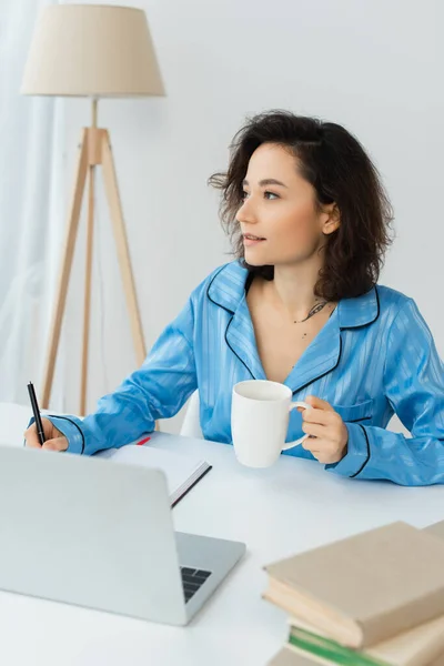 Pensive freelancer holding cup of coffee near laptop on desk — Stock Photo
