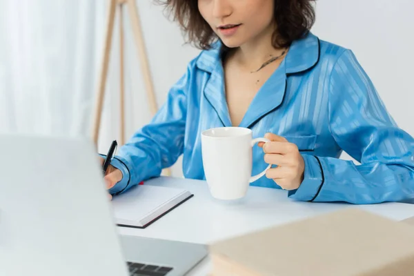 Cropped view of young woman holding cup of coffee near notebook and laptop — Stock Photo