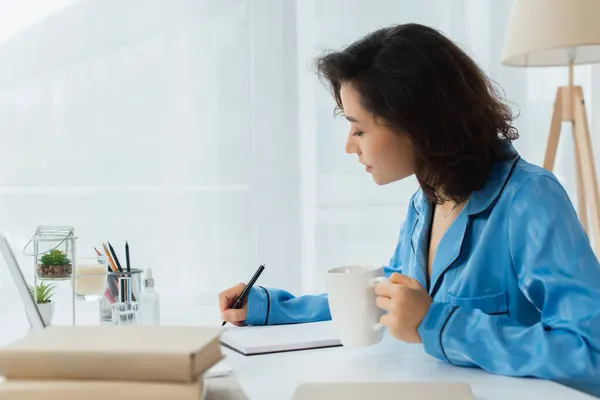 Junge Frau schreibt in Notizbuch, während sie eine Tasse Kaffee in der Hand hält — Stockfoto