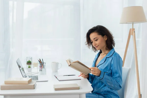 Jeune femme lecture livre près ordinateur portable sur le bureau — Photo de stock