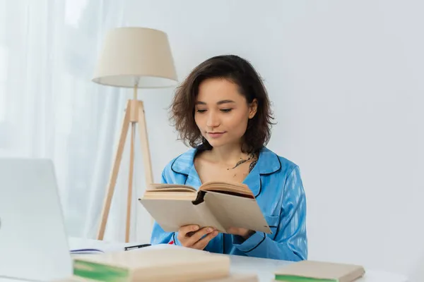 Tatuado jovem mulher leitura livro perto laptop na mesa — Fotografia de Stock