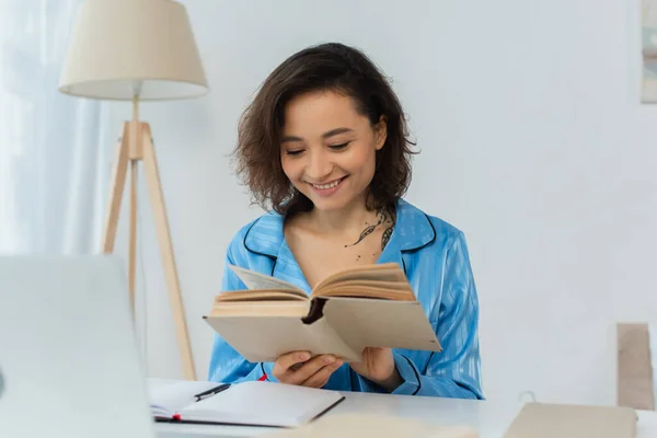 Happy young woman reading book near laptop on desk — Stock Photo