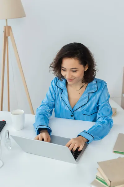 Jovem freelancer alegre usando laptop perto de copo e livros — Fotografia de Stock