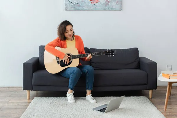 Feliz joven aprendiendo a tocar la guitarra acústica cerca de la computadora portátil en la alfombra - foto de stock