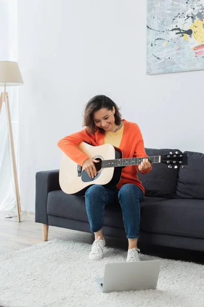 Веселая молодая женщина учится играть на акустической гитаре рядом с ноутбуком на ковре — стоковое фото