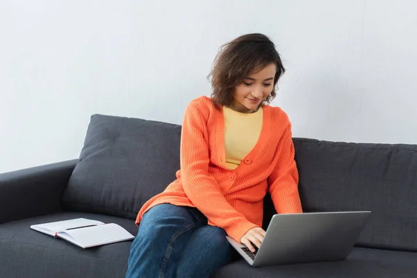 Mujer alegre sentada en el sofá y el uso de la computadora portátil cerca del portátil - foto de stock