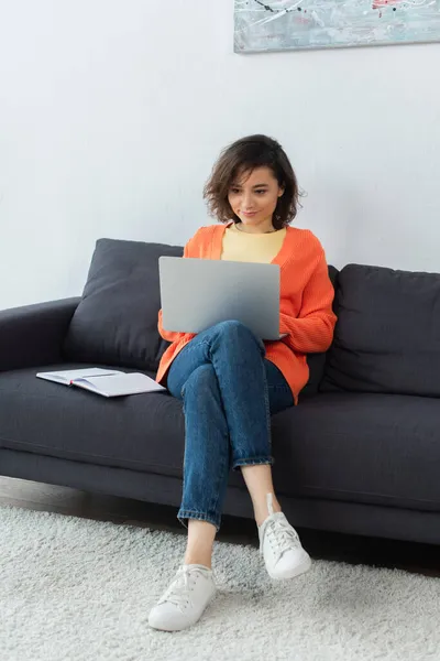 Brunette young woman using laptop near notebook on couch — Stock Photo