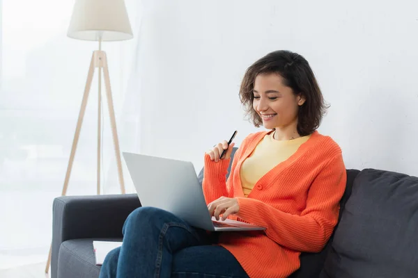 Fröhliche Frau sitzt auf der Couch und hält Stift, während sie Laptop benutzt — Stockfoto