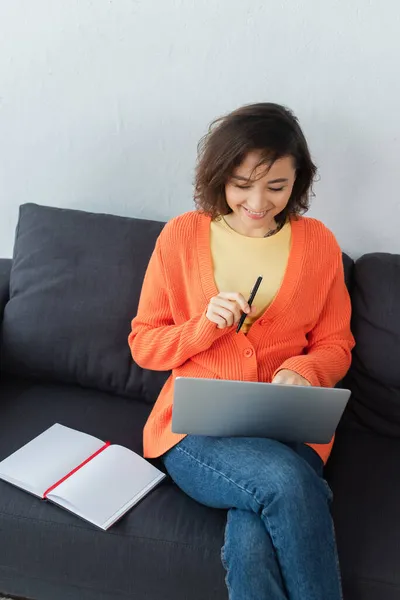 High angle view of cheerful woman sitting on couch and using laptop near notebook — Stock Photo