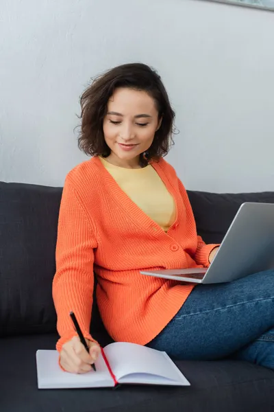 Cheerful woman writing in notebook while using laptop in living room — Stock Photo