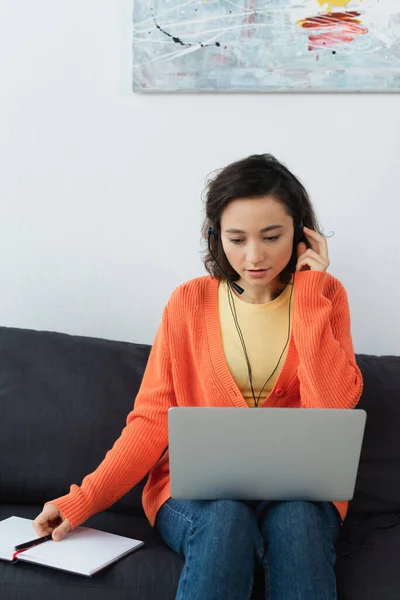 Young operator in headset reaching pen and using laptop in living room — Stock Photo