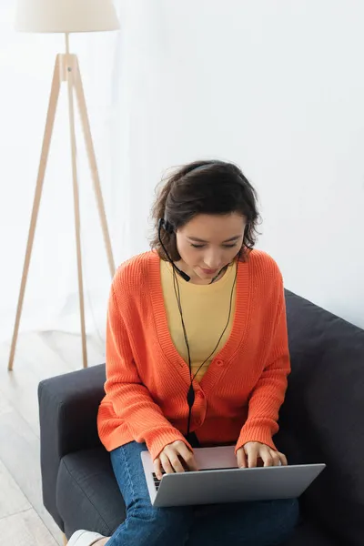 Morena joven operador en auriculares utilizando el ordenador portátil en el sofá - foto de stock