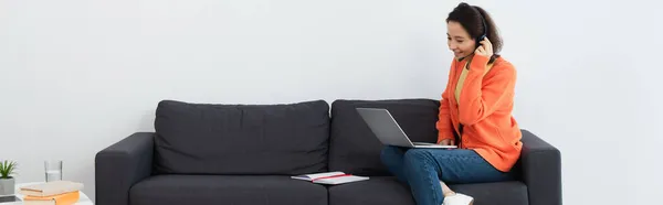 Cheerful operator in headset using laptop while sitting on sofa, banner — Stock Photo