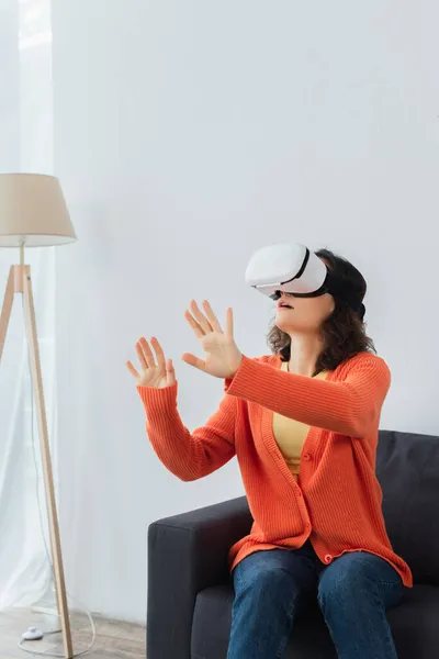 Junge brünette Frau in vr headset gestikulierend beim Spielen und Sitzen auf dem Sofa — Stockfoto