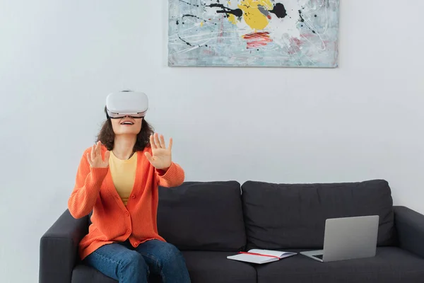 Счастливая женщина в гарнитуре VR жестикулирует, сидя рядом с ноутбуком на диване — стоковое фото