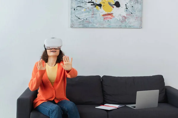 Glückliche Frau in vr Headset gestikuliert, während sie neben Laptop auf Sofa sitzt — Stockfoto