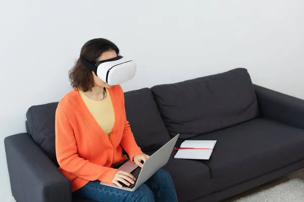 Jeune femme brune dans vr casque à l'aide d'un ordinateur portable tout en étant assis sur le canapé — Photo de stock