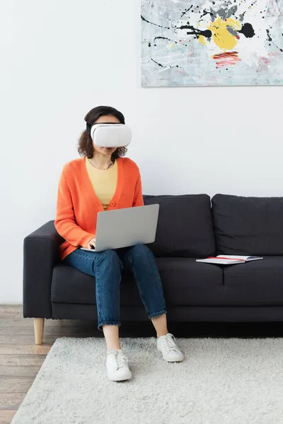 Joven morena mujer en vr auriculares usando el ordenador portátil mientras está sentado en el sofá - foto de stock