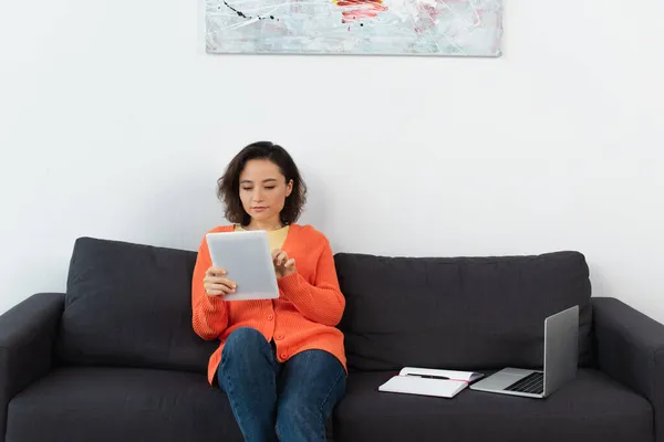 Mujer joven usando tableta digital y sentado cerca de la computadora portátil y portátil en el sofá - foto de stock