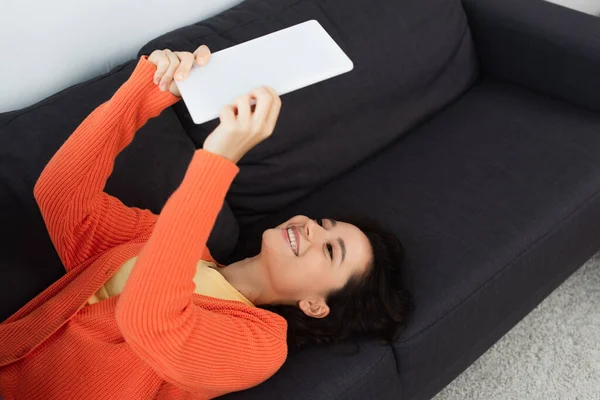 Hochwinkelaufnahme einer glücklichen jungen Frau, die ein digitales Tablet benutzt und auf dem Sofa liegt — Stockfoto