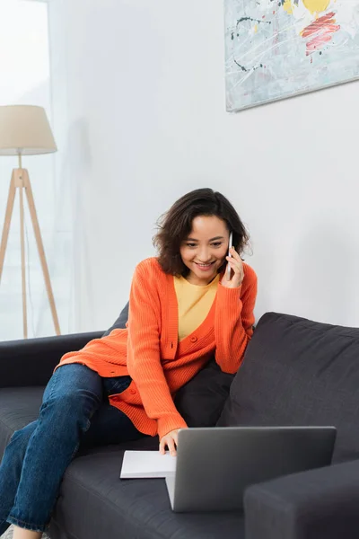 Jovem feliz falando no celular e usando laptop no sofá — Fotografia de Stock