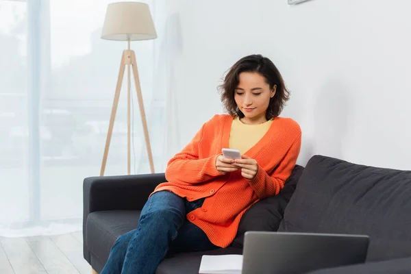 Junge Frau textet auf Handy in der Nähe von Laptop im Wohnzimmer — Stockfoto