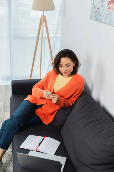 Mujer joven sosteniendo el teléfono inteligente y mirando a la computadora portátil en la sala de estar - foto de stock