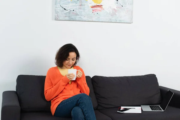 Jovem mulher segurando xícara de café perto de gadgets com tela em branco no sofá — Fotografia de Stock