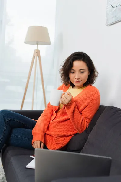 Young freelancer holding cup and looking at laptop on couch — Stock Photo