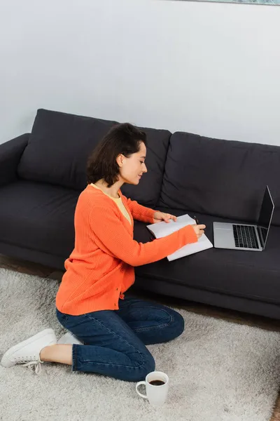 Junge Frau sitzt auf Teppich und schreibt in Notizbuch neben Laptop mit leerem Bildschirm auf Sofa — Stockfoto