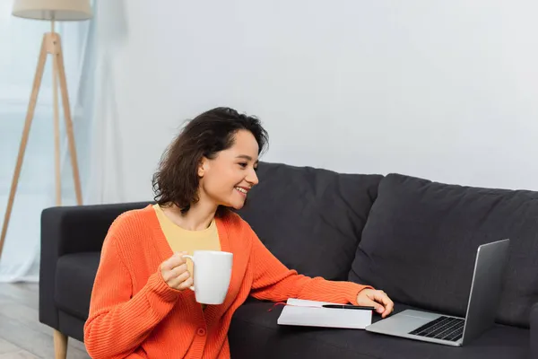 Feliz joven mujer sosteniendo la taza de café mientras mira el ordenador portátil - foto de stock