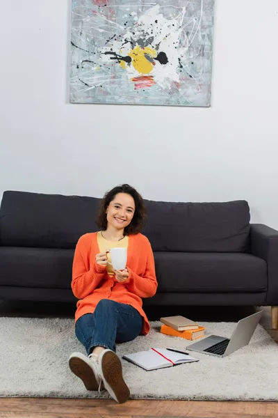 Felice giovane donna seduta su tappeto e con in mano una tazza di caffè vicino a laptop e libri — Foto stock