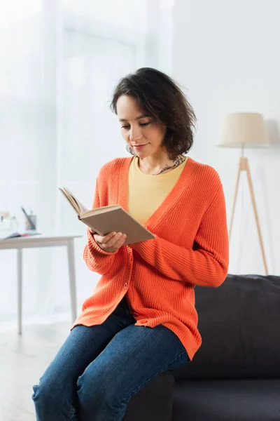 Tatuato donna lettura libro mentre seduto sul divano a casa — Foto stock