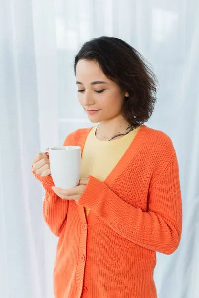 Mujer joven tatuada sosteniendo taza cerca de la cortina blanca - foto de stock