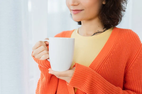 Vista recortada de mujer joven tatuada sosteniendo taza cerca de la cortina blanca - foto de stock