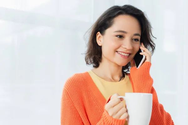 Mujer joven positiva sosteniendo taza y hablando en el teléfono inteligente cerca de la cortina blanca - foto de stock