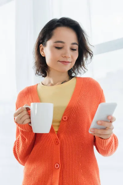 Tätowierte junge Frau mit Tasse und Smartphone nahe weißem Vorhang — Stockfoto