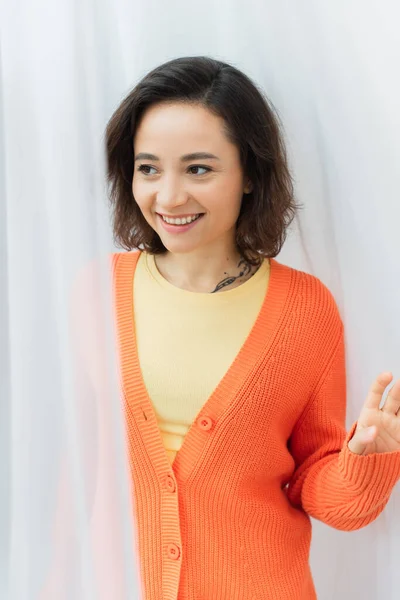 Portrait of happy young woman with tattoo smiling near white curtain — Stock Photo