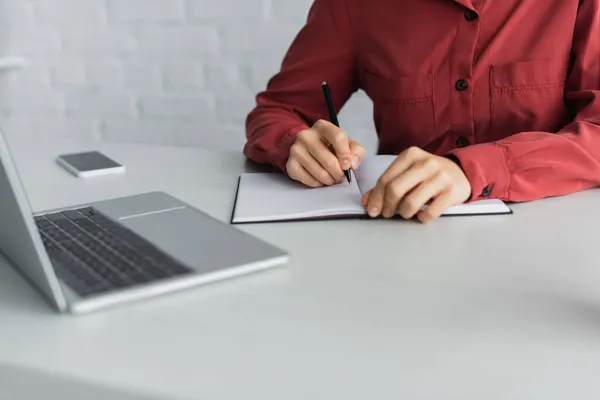 Vista ritagliata dell'insegnante che scrive nel taccuino vicino ai dispositivi sulla scrivania — Foto stock