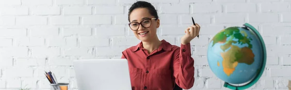 Happy teacher in eyeglasses using laptop and holding pen while working from home, banner — Stock Photo