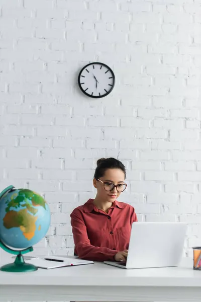 Young teacher in eyeglasses using laptop near globe while working from home — Stock Photo