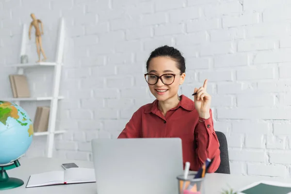 Happy teacher in eyeglasses pointing with finger during video call and online lesson — Stock Photo