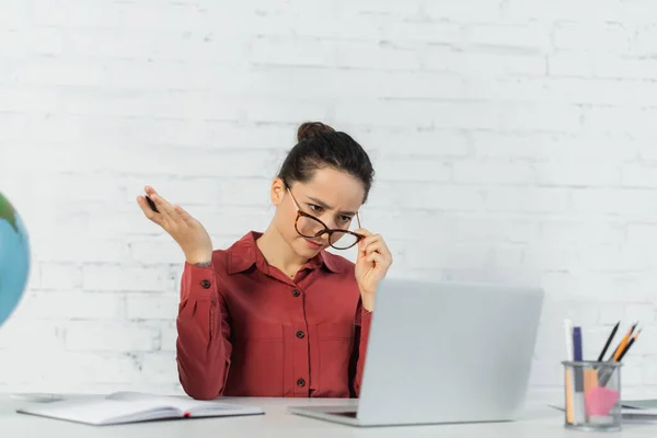 Teacher adjusting eyeglasses while looking at laptop and holding pen — Stock Photo