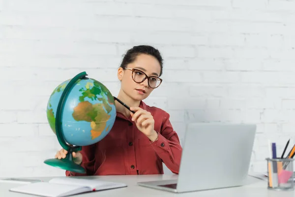 Young teacher in eyeglasses holding pen near globe and looking at laptop — Stock Photo