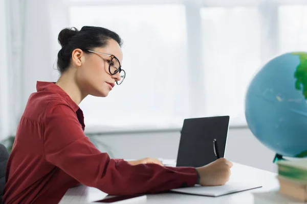 Young teacher in eyeglasses writing in notebook near laptop with blank screen and globe — Stock Photo
