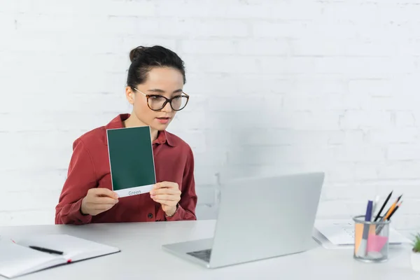 Junge Lehrerin mit Brille hält Karte mit grünem Schriftzug in der Hand und blickt auf Laptop auf Schreibtisch — Stockfoto