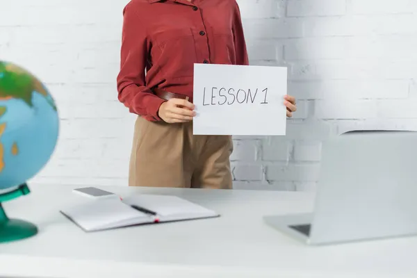 Cropped view of teacher holding paper with lesson one lettering near globe near devices on desk — Stock Photo