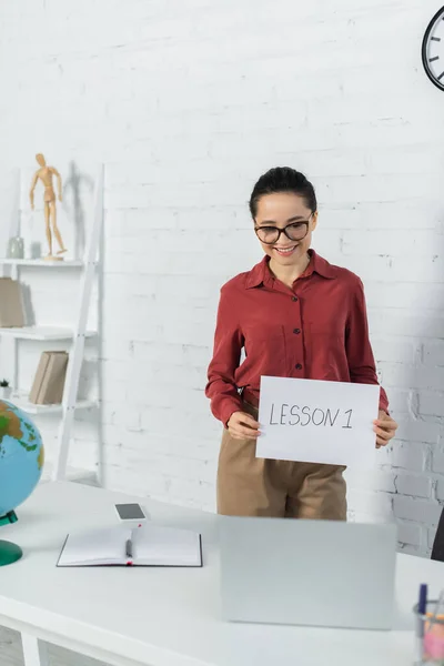 Smiling teacher in eyeglasses holding paper with lesson one lettering and looking at laptop — Stock Photo